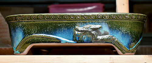 Gyozan – Rechteckige Bonsai-Schale mit gerundeten Ecken, stilisierten Nietenköpfen am oberen Rand und einem geschwungenen Band mit Drachenkopf im unteren Bereich. Diese Schale wurde 2018 von Gyozan getöpfert. Die grüne Oribe-Glasur hat faszinierende Farbverläufe